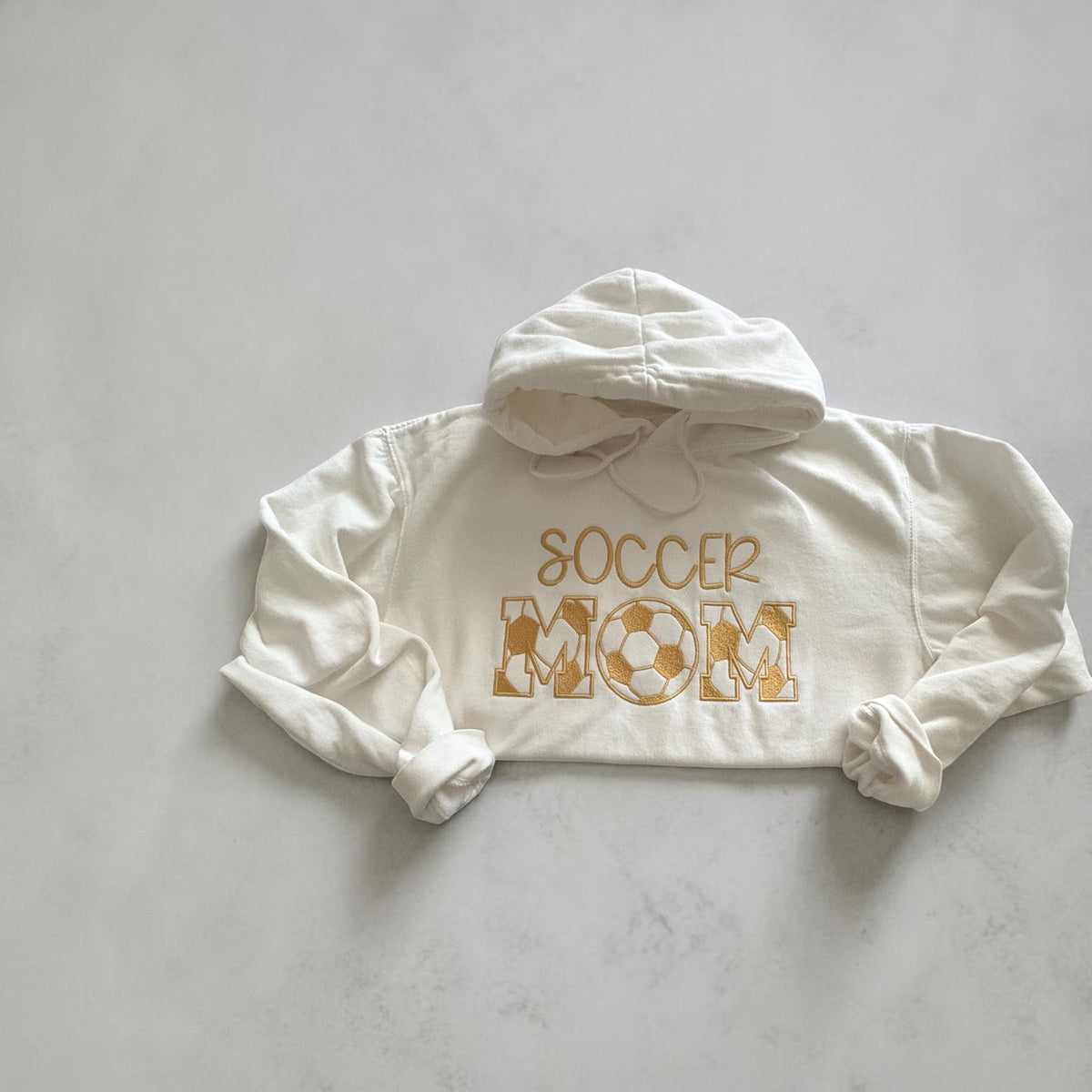 Ivory hoodie with gold Soccer Mom embroidery and a soccer ball forming the O in Mom