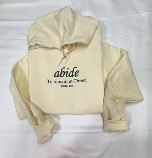Pale yellow hoodie with dark lettering reading abide, To remain in Christ, John 15:4