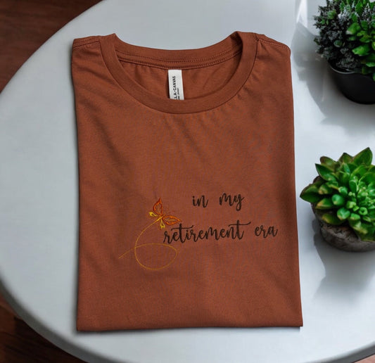 In My Retirement Era embroidered rust T-shirt photographed on a white table with plants